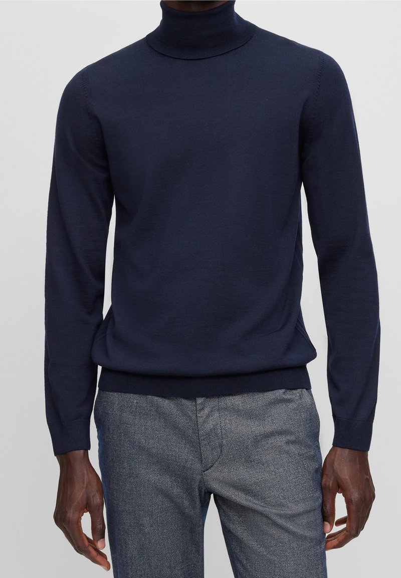 Homme portant un pull à col roulé bleu marine et un pantalon gris texturé sur un fond uni.