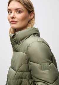 Vadderad jacka i olivgrön, med hög krage, tryckknappar och en texturerad yta med en logotyp-patch på ärmen.