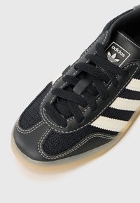 Zwarte Adidas-sneaker met witte strepen, met een gestructureerde bovenkant, veters en een rubberen zool, geplaatst tegen een lichtgrijze achtergrond.