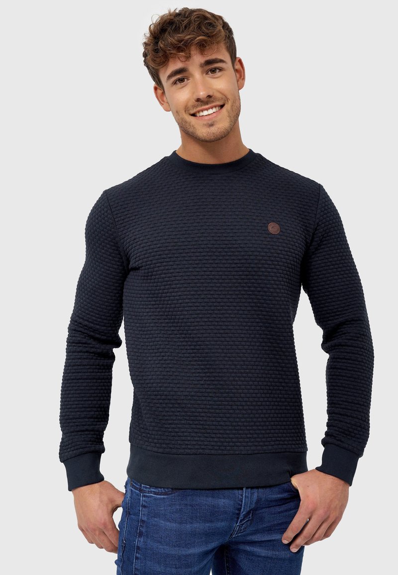 Maglione blu navy con design in tessuto testurizzato, scollo rotondo, polsini e orlo a coste, con una piccola toppa del logo marrone sul petto.