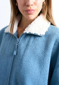 Femme portant une veste en polaire bleue avec un col sherpa blanc, fermeture éclair partiellement fermée, ne montrant que les lèvres et le menton.