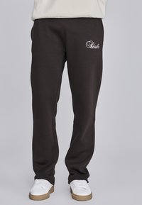 Zwarte sweatpants met geborduurd wit logo, elastische tailleband en taps toelopende broekspijpen. Zachte stof met een gladde textuur. Gecombineerd met witte sneakers.