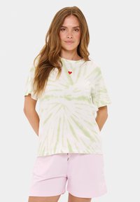 Balta marškinėlių su žaliais tie-dye raštais, apvaliu iškirpimu, trumpomis rankovėmis ir raudono širdies siuvinėjimu krūtinėje. Suderinti su rožiniais šortais.