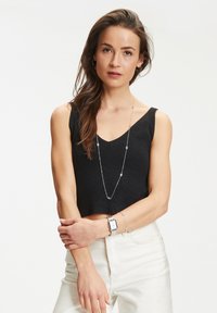 Schwarzes geripptes Crop-Top mit V-Ausschnitt, kombiniert mit weißen hochgeschnittenen Jeans. Die Accessoires umfassen eine silberne Halskette und eine Uhr.