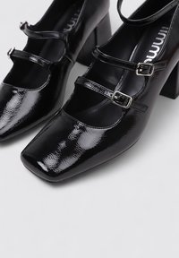 Zapatos de tacón alto negros con un acabado brillante, puntera cuadrada, dos tiras con hebillas y un tacón ancho. Textura suave en toda la superficie.