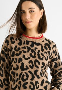 Beiger Pullover mit schwarzem Leopardenmuster und einem gerippten roten Kragen. Weicher Stoff, lässige Passform und lange Ärmel. Akzent durch eine goldene Halskette.