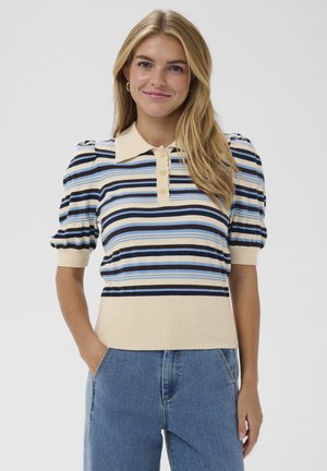 Mladá žena s dlhými blond vlasmi, oblečená v modrom, čiernom a béžovom pruhovanom svetríku s krátkym rukávom a modrých džínsoch, usmievajúca sa s jednou rukou v vrecku.