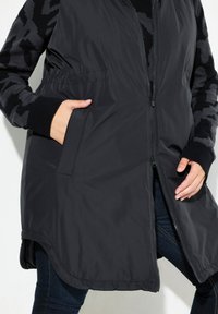 Schwarze West mit einem Reißverschluss vorne, die eine glatte Oberfläche und kontrastierende Ärmel im Camouflage-Muster aufweist. Enthält Seitentaschen.