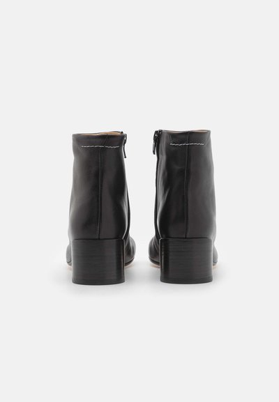 Botas de tornozelo em couro preto com textura suave, fecho lateral com zíper e um salto de madeira empilhado. Design minimalista com detalhes de costura branca.