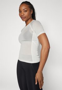 Weißes kurzärmliges Mesh-Shirt mit figurbetontem Design und strukturiertem Stoff. Mit kontrastierenden Nähten und rundem Ausschnitt, kombiniert mit schwarzen Leggings.