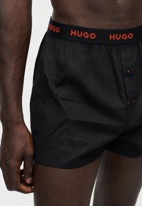Personne portant un boxer Hugo noir avec une ceinture noire et un logo Hugo rouge, vue de la taille jusqu’au milieu de la cuisse.