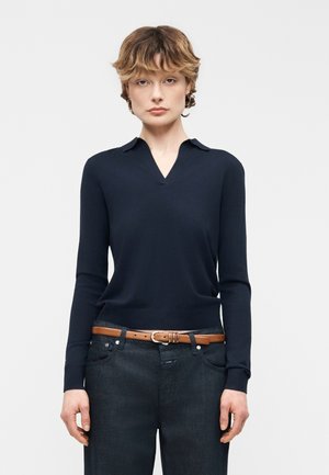 Kvinde med kort hår iført en mørkeblå langærmet skjorte med krave, stoppet ned i mørke jeans med et brunt bælte, stående mod en ensfarvet baggrund.