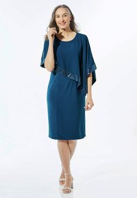 Teal jurk met een vloeiende cape-achtige mouw met sequins langs de zoom. Gladde stof, knie-lengte, gecombineerd met lichte sandalen.