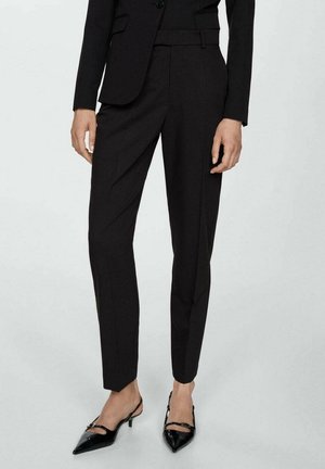 Woman wearing black tailored trousers, black blazer, and black pointed-toe slingback heels standing against a plain background.