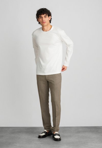 Bílá košile s dlouhým rukávem, slim-fit béžové texturované kalhoty a dvoubarevné černobílé boty. Jednoduché pozadí, model čelí vpřed.