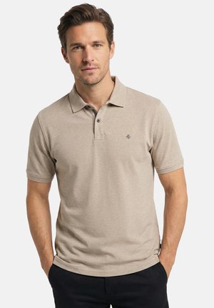 Mand med kort brunt hår iført beige kortærmet polo med lille logo på brystet og sorte bukser, hænderne i lommerne, neutral baggrund.