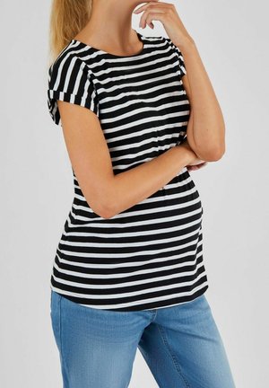 Vrouw in zwart-wit gestreepte korte mouw shirt en blauwe jeans, met gekruiste armen, waarbij ze met één hand haar kin aanraakt tegen een effen achtergrond.