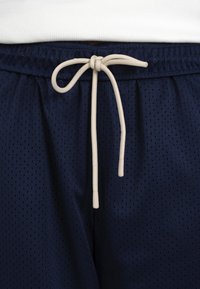 Shorts de sport bleu marine avec cordon blanc noué en nœud au centre de la taille, portés avec un top blanc.