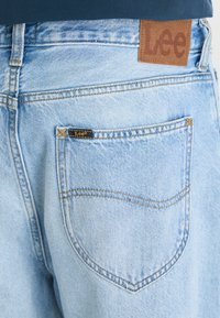 Jeans in denim azzurro chiaro con un patch in pelle marrone con la scritta "Lee" in rilievo. La tasca posteriore ha un'etichetta nera con "Lee" e cuciture visibili.