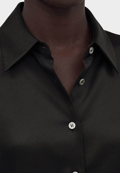 Κοντινό πλάνο ενός σκούρου δέρματος ατόμου που φοράει μαύρο πουκάμισο με μύτη γιακά και λευκά κουμπιά, σε απλό φόντο.