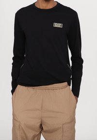 Černé dlouhé rukávy tričko s kulatým výstřihem a logem ve zlaté barvě; spárováno se béžovými kalhotami s elastickým pasem a bočními kapsami.