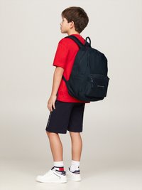 Svart ryggsäck med dragkedjor och framficka, bärs av ett barn i en röd t-shirt och marinblå shorts, placerad mot en neutral bakgrund.