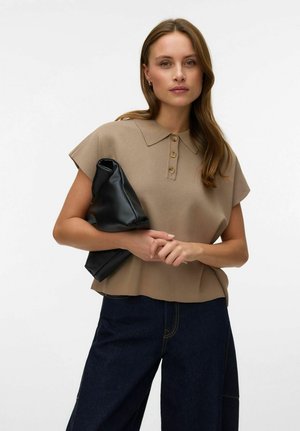 Naine pikkade juustega, kellel on seljas beež lühikeste varrukatega kraega särk ja tumedad teksad, hoides volditud musta nahast väljapilu.