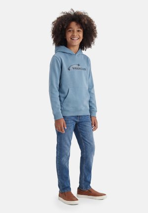 Laps lokkis juustega, kandes sinist Wrangleri dressipluusi, siniseid teksapükse ja pruune libisevaid kingi, naeratav ja seismas valge tausta ees.