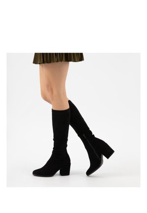 Jambes portant des bottes en daim noires montantes jusqu'aux genoux avec des talons épais et des fermetures éclair latérales, associées à une jupe plissée courte vert olive.