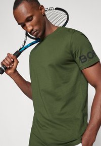 Grønn bomullst-skjorte med rund hals og korte ermer, med en svart "BOSS" logo på venstre arm. Holder en tennisracket.