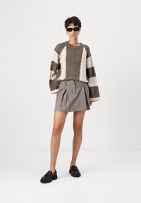 Maglione lavorato a maglia a righe marroni e crema con motivi a treccia. Abbinato a una gonna plissettata a quadri grigio e mocassini neri. Indossati occhiali da sole neri.