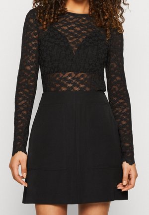 Femme portant un haut à manches longues en dentelle florale noire associé à une jupe noire taille haute avec poches avant.
