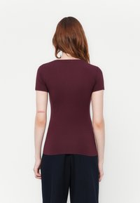 Donna con lunghi capelli castani che indossa una maglietta attillata a maniche corte di colore bordeaux e pantaloni neri larghi, in piedi con la schiena rivolta in avanti.