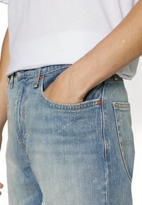 Light blue denim jeans with a relaxed fit, five pockets, and faded areas. Includes copper rivets and stitching details.