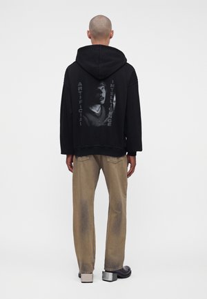 Homme portant un sweat à capuche noir avec un graphisme et texte "Artificial Intelligence" dans le dos, pantalon beige et chaussures noires à talons argentés, debout de dos.