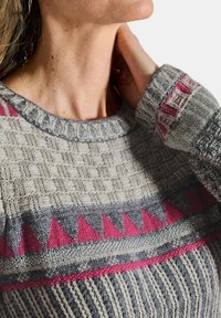 Nahaufnahme einer Person, die einen grauen Strickpullover mit pinken und dunkelgrauen geometrischen Mustern trägt, während sie mit der Hand den Hals berührt.