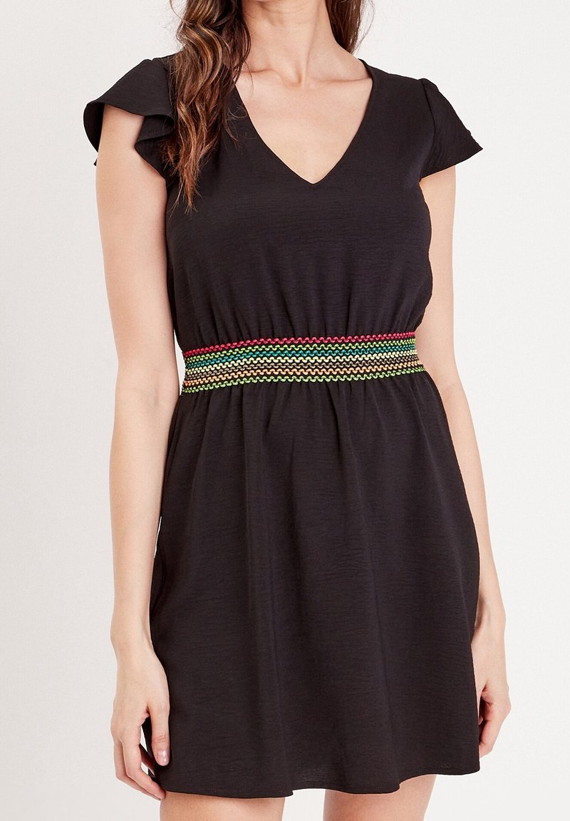 Femme portant une robe noire courte avec des manches volantées et une ceinture tissée multicolore à la taille, debout devant un fond uni.