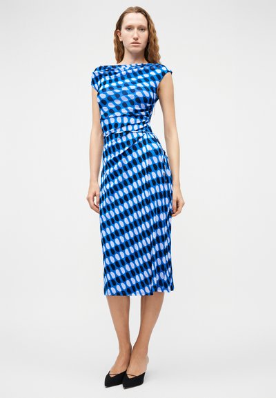 Γυναίκα που φορά μια μεσομήκη μπλε φόρεμα με μοτίβα και κοντά μανίκια, συνδυασμένη με μαύρα μυτερά τακούνια, στέκεται μπροστά σε απλό λευκό φόντο.