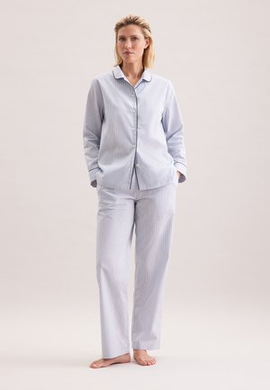 Vrouw staat blootsvoets en draagt een lichtblauw, lang mouwen, met knopen pyjamashirt en bijpassende rechte pyjamabroek tegen een neutrale achtergrond.