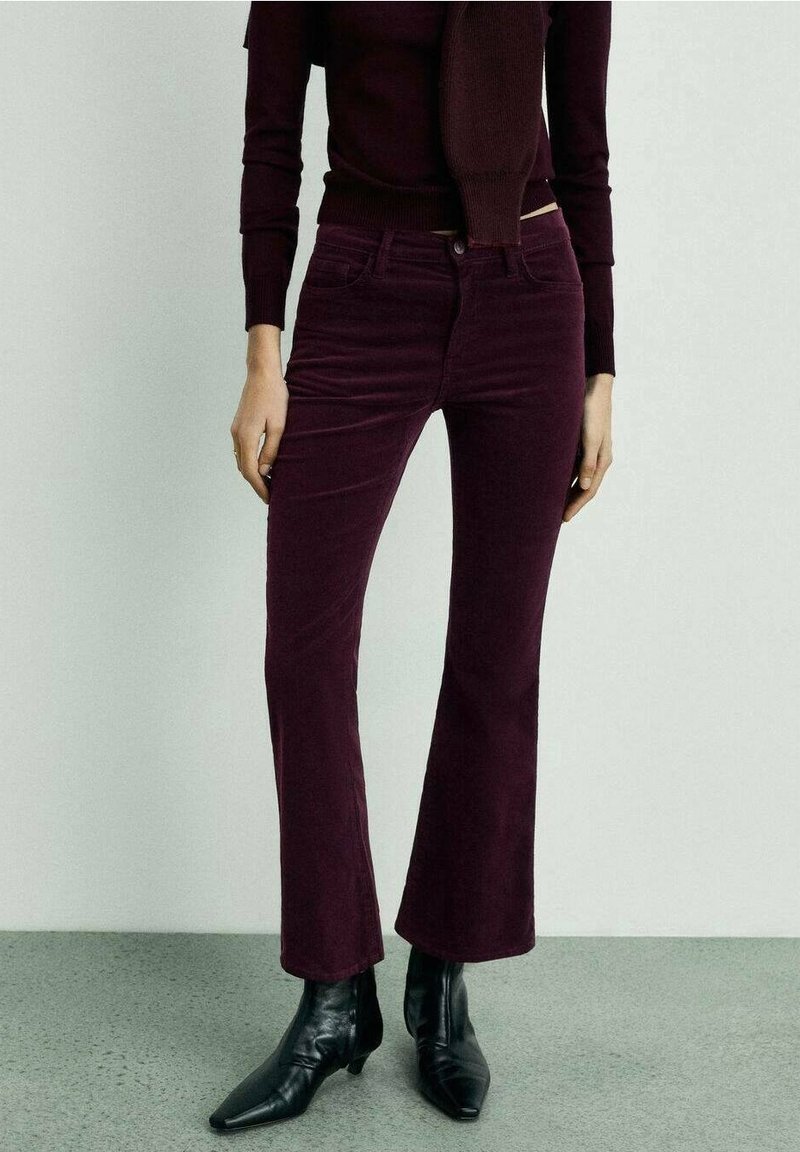 Femme portant un pantalon évasé ajusté violet foncé, un haut assorti et des bottines noires à bout pointu sur un fond clair uni.