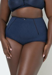 Kobieta w większym rozmiarze ubrana w ciemnoniebieską bieliznę z wysokim stanem i pasujący biustonosz, ręce spoczywają na udach na jednolitym tle.