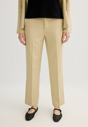 Pantalones beige a medida combinados con bailarinas Mary Jane negras tejidas y una parte superior negra en una persona de pie.