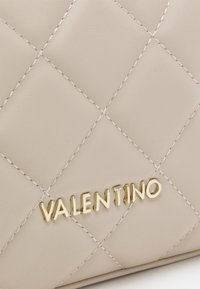 Pamušaluotas smėlio spalvos odinis rankinis krepšys su auksiniu "Valentino" logotipu, matomomis siūlėmis ir minkšta tekstūra.