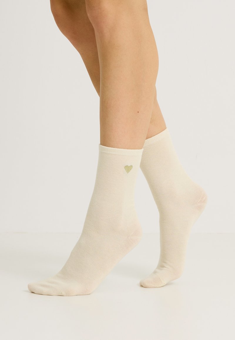 Calcetines tobilleros de color beige, hechos de un material suave, que presentan un pequeño diseño de corazón verde en el lado exterior. Textura suave y puño acanalado.