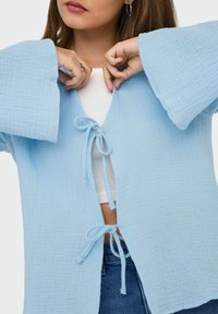 Giacca azzurro chiaro, morbida e ampia con maniche larghe, tessuto testurizzato e chiusura anteriore con nodi. Indossata sopra una maglietta bianca e abbinata a jeans.