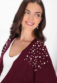 Burgundowy kardigan o prążkowanej fakturze, z rozsianymi sztucznymi perłami na ramionach. Nałożony na biały top z dekoltem w serek.