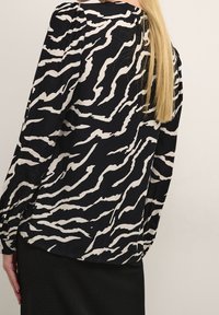 Blouse en imprimé zèbre noir et blanc avec des manches longues, un col rond et des détails plissés aux épaules. Tissu doux, coupe loose et ourlet arrondi.