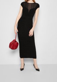Robe noire ajustée avec des manchons, décolleté en mesh transparent, texture côtelée verticale, assortie à un sac à main rouge et des talons noirs.