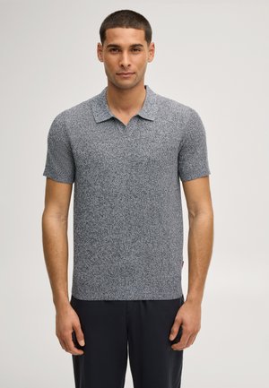 Uomo con capelli corti che indossa una polo corta a maniche corte di maglia grigia strutturata e pantaloni neri su uno sfondo chiaro e uniforme.