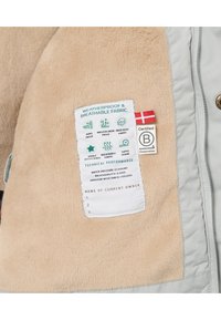Beige fleeceinteriør med grå værbestandig utside. Merket viser stoffegenskaper: vindbestandig, vannavvisende, pustende, med ytelsesdetaljer.